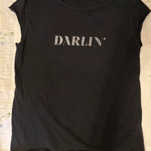 Svart t-shirt med glittrig text från NA-KD - Snygg svart t-shirt från NA-KD med text 'DARLIN' på framsidan. Perfekt för en casual look med en touch av glamour. Kortärmad och bekväm passform.