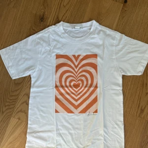 Vit t-shirt med hjärtmönster från Monki - Säljer en vit t-shirt från Monki med ett orange hjärtmönster på framsidan. T-shirten är kortärmad och gjord i bomull, perfekt för en avslappnad stil nu när sommaren kommer såklart stryks den innan den blir skickad.