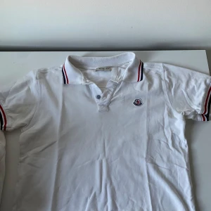 Vit pikétröja från Moncler - Säljer en stilren vit pikétröja från Moncler med klassisk krage och knappar. Tröjan har korta ärmar med röd och blå randdetalj på krage och ärmslut. Perfekt för en avslappnad men snygg look.   Ej använd , pris kan diskuteras privat 