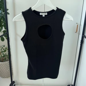 Svart ärmlös topp från Saemdi - Snygg svart ärmlös topp från Saemdi med en unik cut-out detalj framtill. Toppen är ribbad och har en tight passform, perfekt för en trendig look. Passar bra till både jeans och kjol!