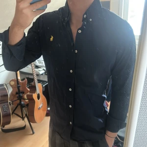 Mörkblå skjorta med gul broderad logga - Snygg mörkblå skjorta från Ralph Lauren med en gul broderad logga på bröstet. Skjortan har långa ärmar och knappar framtill. Perfekt för en stilren look.