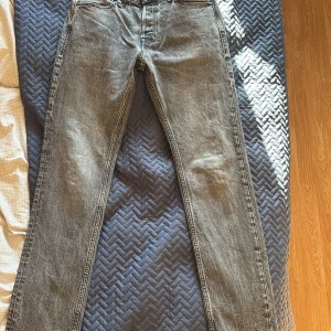 Grå jeans från Jack & Jones - Säljer ett par grå jeans från Jack & Jones. De har en klassisk design med fem fickor och en dragkedja. Perfekta för en avslappnad stil. Märket är känt för sin kvalitet och stilrena plagg.