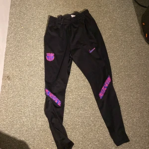Svarta träningsbyxor från Nike - Säljer ett par svarta träningsbyxor från Nike med Dri-FIT-teknologi. Byxorna har en elastisk midja och är dekorerade med ett lila Nike-märke och ett klubbemblem. Perfekta för fotbollsträning eller annan sportaktivitet.
