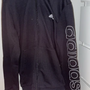 Svart hoodie från Adidas - Säljer en svart hoodie från Adidas med dragkedja och stor logga längs ärmen. Perfekt för en sportig look eller avslappnad stil. Den har en bekväm passform och huva för extra komfort.