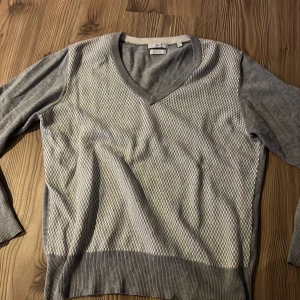 Grå v-ringad tröja - Säljer en stilren grå tröja med v-ringning och ett diskret rutmönster på framsidan. Tröjan har långa ärmar och ribbade kanter vid ärmslut och nederkant. Perfekt för en avslappnad men ändå snygg look.