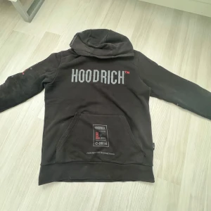 Svart hoodie från Hoodrich - Säljer en svart hoodie från Hoodrich med tryck både på ryggen och ärmen. Hoodien har en stor ficka framtill och en justerbar huva. Perfekt för en avslappnad stil.