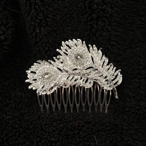 Silverfärgad hårkam med strass - Elegant hårkam i silverfärg med glittrande strassdetaljer formade som påfågelfjädrar. Perfekt för att ge en extra touch av glamour till din frisyr.
