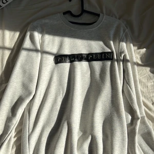 ÄKTA Grå tröja från Philipp Plein - Säljer en stilren grå tröja från Philipp Plein med ett svart märke på bröstet och en liten logga på ryggen. Tröjan har långa ärmar och en rund halsringning. Perfekt för en avslappnad look.