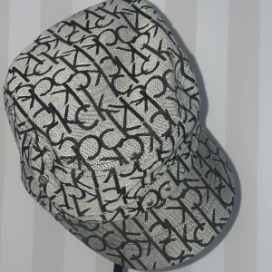 Mönstrad keps från Calvin Klein - Snygg keps från Calvin Klein med ett stilrent mönster av CK-loggor i svart på en grå bakgrund. Kepsen har en klassisk form och är tillverkad i en blandning av polyester och bomull. Perfekt för att ge din outfit en trendig touch. Mått runtom 60 cm.