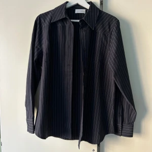 Mörkblå randig skjorta från Carin Wester - Snygg mörkblå skjorta från Carin Wester med vita ränder. Skjortan har långa ärmar och knappar framtill. Perfekt för en stilren look. Aldrig använd. 