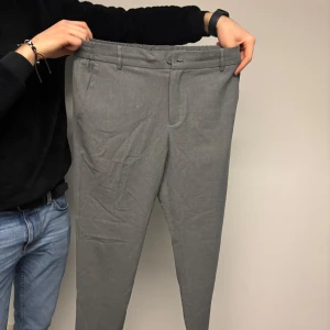 Grå kostymbyxor från Les Deux - Snygga grå kostymbyxor från Les Deux med en klassisk design. Byxorna har en normal passform och är perfekta för en stilren look. De har en dragkedja och knapp framtill för enkel stängning.