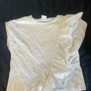 Vit topp med volangdetalj från Zara - Säljer en söt vit topp från Zara storlek 152  med volangdetalj på framsidan. Toppen har en rund hals och är kortärmad, perfekt för varmare dagar. Passar både till jeans och kjol för en avslappnad look.