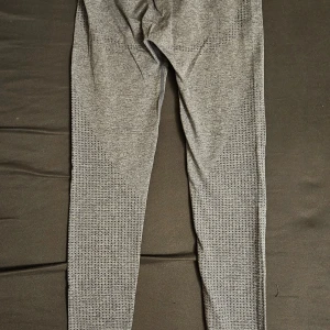 Grå träningstights från Gymshark - Säljer ett par grå träningstights från Gymshark med diskret mönster längs benen och logga på höften och vristen. Tightsen har hög midja och är tillverkade i ett stretchigt material som passar perfekt för gym eller löpning.