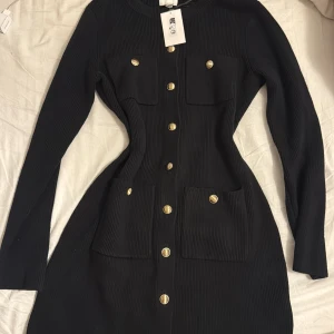Svart ribbad klänning med guldknappar - Snygg svart ribbad klänning med långa ärmar och guldiga knappar framtill. Klänningen har fyra fickor och markerad midja för en smickrande siluett. Helt ny med prislapp 