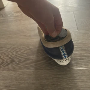 Blå sneakers från The Gallerist med vit klöverdetalj - Säljer ett par blå sneakers från The Gallerist med vit klöverdetalj på sidan och vit sula. Skorna har snörning och en dekorativ mönstrad remsa baktill. Perfekta för dig som vill sticka ut med en unik design.