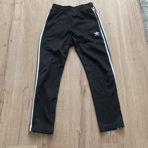Svarta träningsbyxor från Adidas med vita ränder - Svarta Adidas träningsbyxor med klassiska vita ränder längs sidorna och broderad logga på ena benet. Byxorna har resår i midjan och är tillverkade i Primeblue-material som innehåller återvunnen plast från havet.