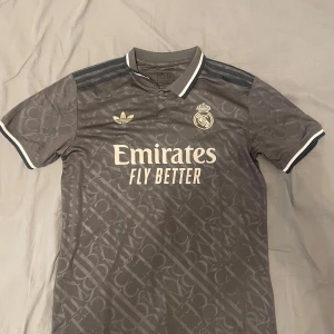 Grå Real Madrid fotbollströja från Adidas - Säljer en grå Real Madrid fotbollströja från Adidas med klubbmärke och sponsortryck på bröstet. Tröjan har korta ärmar, vit krage och detaljer samt diskret mönster i tyget. Perfekt för dig som älskar fotboll och vill visa ditt stöd för Real Madrid.