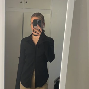 Svart skjorta med klassisk krage - Säljer en svart skjorta med klassisk krage och knäppning framtill. Skjortan har lång ärm och en rak passform som ger en stilren look. Perfekt att styla till både jeans och kostymbyxor.