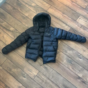 Svart dunjacka med huva från Peak Performance - Säljer en svart dunjacka från Peak Performance med huva och dragkedja. Jackan är vadderad och har en klassisk pufferstil som håller dig varm under kalla dagar. Perfekt för dig som vill ha en stilren och funktionell jacka.