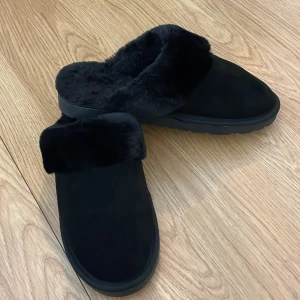 Svarta tofflor med fuskpäls - Mysiga svarta tofflor med mjuk fuskpäls runt öppningen och insidan. Perfekta att glida runt i hemma under kyliga dagar. Yttermaterialet är i mocka. Tofflorna är aldrig använda. folk med storlek 39 kan ha dom.