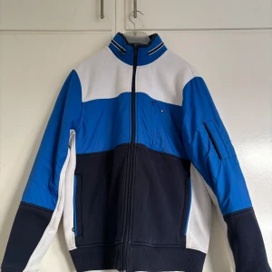 Blå och vit jacka från Tommy Hilfiger - Snygg jacka från Tommy Hilfiger i blått, vitt och svart. Jackan har hög krage, dragkedja framtill och två fickor med dragkedja. Perfekt för dig som gillar sportig stil och vill sticka ut med klassiska färger.