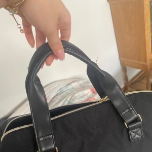 Svart weekendväska från BOSS Parfums - Snygg svart weekendväska från BOSS Parfums med dubbla handtag i skinnimitation och guldfärgad dragkedja. Väskan har en stilren design med BOSS-logga framtill och detaljer i syntetmaterial. Perfekt för resor eller gymmet.