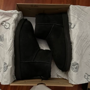 Svarta UGG Classic Mini boots - Säljer ett par svarta UGG Classic Mini boots i mocka. Skorna har en låg skaftmodell och är fodrade för extra värme. Perfekta för kyliga dagar och har en klassisk rund tå samt platt sula.