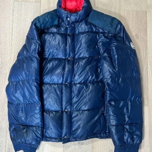 vintage moncler jacka  - Snygg blå pufferjacka från Moncler med röd insida och klassisk logga på ärmen. Jackan har hög krage, knappar och dragkedja framtill. Perfekt för kyliga dagar när du vill hålla stilen.