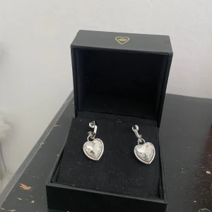 Silverfärgade hjärtformade örhängen med kristall - Liknar Maria Nilsdotter🩷 Säljer ett par silverfärgade örhängen med hjärtformade hängen och gnistrande kristall på framsidan. Örhängena har en modern, kantig båge och stift. Perfekta för dig som vill ha något extra till din stil.