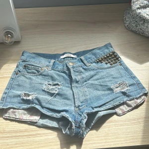 Jeansshorts med nitar och slitningar - Snygga blå jeansshorts från Ivyrevel!💙De har slitningar, blommiga innerfickor och snygga guldiga nitar på ena fickan. Passar perfekt t.ex. med en bikini eller stickad tröja!👙💕   Midjemått rakt över: 37-38cm
