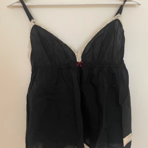 Svart linnetopp med spets från Odd Molly - Svart linnetopp från Odd Molly med justerbara axelband och dekorativ vit spets längs kanterna. Liten rosett framtill och broderad logga på ryggen. Lappen nertill har texten 'celebrating yourselves'. Perfekt för lager-på-lager-stil.