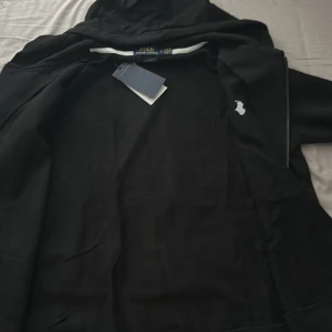 Svart hoodie från Polo Ralph Lauren - Svart hoodie från Polo Ralph Lauren med dragkedja och huva. Klassisk design med vit broderad logga på bröstet och ribbade muddar. Perfekt för en avslappnad och stilren look.