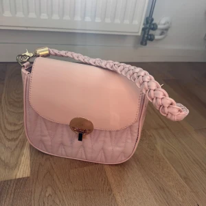 Ljusrosa quiltad axelväska med flätat axelband - Säljer en söt ljusrosa axelväska med quiltad framsida och ett flätat axelband. Väskan har ett guldfärgat spänne framtill och är perfekt för att lyfta vilken outfit som helst.