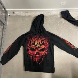 Svart hoodie med eldsflammor och dödskalle från Spiral - Säljer en svart hoodie från Spiral med stort tryck av en dödskalle i röd och orange eld på bröstet och flames längs ärmarna. Tröjan har huva och magficka. Perfekt för dig som gillar alternativa och unika plagg.