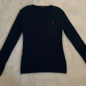 Mörkblå stickad tröja från Polo Ralph Lauren - Säljer en mörkblå stickad tröja från Polo Ralph Lauren med klassisk röd broderad logga på bröstet. Tröjan har lång ärm och smal passform med ribbad struktur. Perfekt för en stilren och tidlös look.