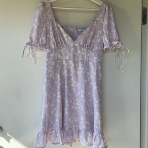 Ljuslila blommig klänning från Loavies - Supersöt ljuslila klänning från Loavies med vita och gula blommor. Klänningen har korta puffärmar med knyt, v-ringning, volangkant nertill och dragkedja i sidan. Perfekt för vår och sommar!