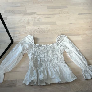 Vit smockad blus från H&M - Säljer en söt vit blus från H&M med smockad överdel och långa puffärmar. Blusen har volang nertill och en romantisk känsla med lätt rynkade detaljer vid axlarna. Perfekt för dig som gillar feminina toppar! Har en liten fläck på framtiden. Går säkert bort om man orkar fixa. 