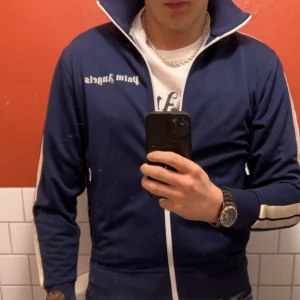 Palm angels track jacket  - Säljer en blå track jacket från Palm Angels med vita/beiga  ränder längs ärmarna och vit dragkedja. Tröjan är i mcyket fint skick har används max 5 gånger. Säljer  för den tyvärr är för liten. Storlek medium vid frågor skicka ett meddelande. 