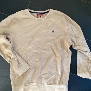 Beige sweatshirt från MCS - Säljer en beige sweatshirt från MCS med rund halsringning och ett litet blått broderat motiv på bröstet. Tröjan har långa ärmar och är perfekt för en avslappnad stil. Klassisk och enkel design.