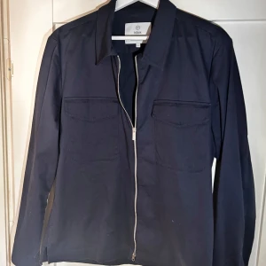 Mörkblå overshirt med dragkedja från Bläck - Säljer en stilren mörkblå overshirt från Bläck med dragkedja framtill och två bröstfickor. Jackan har klassisk krage och lång ärm, perfekt att slänga på sig över en t-shirt för en clean look.