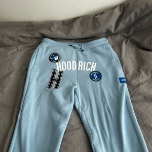 Ljusblå mjukisbyxor från Hoodrich - Säljer ett par ljusblå mjukisbyxor från Hoodrich med broderade patches och logga framtill. Byxorna har dragsko i midjan och flera coola detaljer som ger streetwear-vibe. Perfekta för en avslappnad stil. Helt oandvänd.