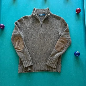 Polo Ralph Lauren Stickad - Säljer en brun stickad tröja från Polo Ralph Lauren med halv dragkedja och förstärkta mockadetaljer på armbågarna. Tröjan har hög krage och långa ärmar, perfekt för kyliga dagar. Riktigt bra skick.