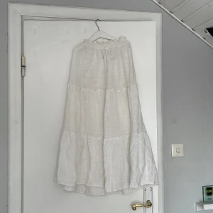 Vit långkjol från H&M - Säljer en luftig och somrig vit långkjol från H&M med volanger och knyt i midjan. Kjolen är tillverkad i ett lätt och skönt tyg, perfekt för varma dagar. Enkel att matcha med olika toppar för en avslappnad stil.