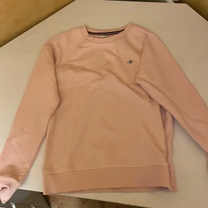 Ljusrosa sweatshirt från Gant - Säljer en ljusrosa sweatshirt från Gant med broderad logga på bröstet. Tröjan har rund halsringning och långa ärmar. Perfekt för en avslappnad och stilren look. Liten i storlek så mer som en 152 cm. 