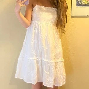 Vit sommarklänning med broderade blommor - Söt vit klänning med tunna axelband och broderade blommor. Klänningen är ärmlös, har en luftig passform och slutar ovanför knäna. Perfekt till studenten! 