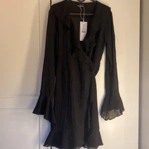 Svart omlottklänning med volang - Säljer en helt ny svart omlottklänning med volangdetaljer och långa ärmar. Klänningen har v-ringning och lätt utställda ärmslut, vilket ger en elegant och trendig look. Perfekt för dig som gillar stilrena plagg med fina detaljer.