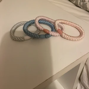 Färgglada flätade armband - Säljer fyra snygga flätade armband i olika färger: vitt, blått, ljusrosa och beige. De är gjorda av syntetmaterial och har en enkel, rund form med matchande plastdetalj. Perfekta som accessoar för att liva upp din outfit!
