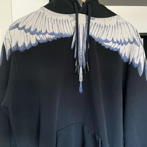 Svart hoodie med vingmönster från Marcelo Burlon - Säljer en svart hoodie från Marcelo Burlon med unikt vingmönster i vitt och blått över axlar och bröst. Tröjan har huva med dragsko och känguruficka framtill. Perfekt statement-plagg för dig som vill sticka ut.