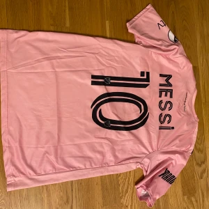 Inter Miami Messi fotbollströja rosa - Säljer en rosa Inter Miami fotbollströja med Messi 10 på ryggen. Tröjan har svarta detaljer, Adidas-logga och sponsortryck på framsidan. Kortärmad modell med klubbmärke på bröstet och tryck på ärmarna.