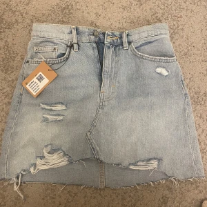 Ljusblå jeanskjol med slitningar - Säljer en ljusblå jeanskjol med råa kanter och slitna detaljer framtill. Kjolen har klassisk femficksdesign och knäppning med knapp och dragkedja. Perfekt för dig som gillar en avslappnad och trendig stil.